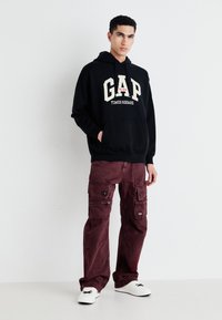 Felpa nera con cappuccio con logo "GAP TIMES SQUARE", abbinata a pantaloni cargo bordeaux con molte tasche e scarpe bianche.