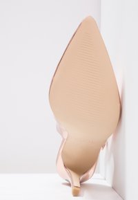 Chaussure à talons aiguilles en or rose avec un bout pointu, finition vernie lisse, semelle texturée et un talon aiguille mince.