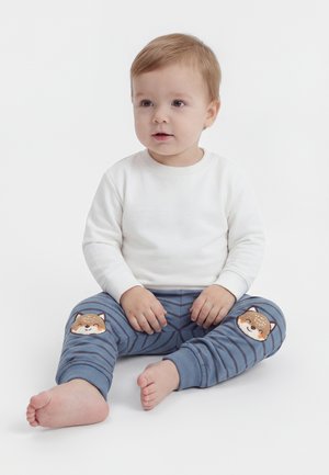 Tout-petit aux cheveux châtain clair assis pieds nus, portant une chemise blanche à manches longues et un pantalon bleu rayé avec des têtes de renard aux genoux.