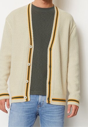Mann trägt einen cremefarbenen gestrickten Cardigan mit gelben und grünen Streifen, dunkelgrünes T-Shirt und hellblaue Jeans, steht vor weißem Hintergrund.