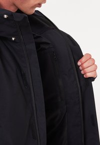 Veste noire avec une poche intérieure zippée, des fermetures à pression et des accents en Velcro. Texture lisse et design moderne avec une silhouette ajustée.