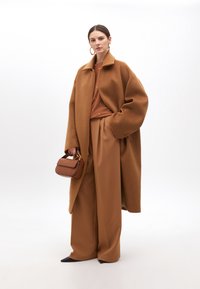 Hnědý vlněný kabát s oversized střihem, kombinovaný s rustikálně zbarveným topem, širokými hnědými kalhotami a malou hnědou kabelkou. Černé boty s špičatou špičkou.