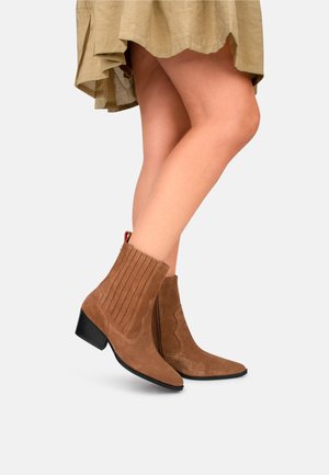 Bottines en daim beige avec un talon bloc, présentant un design à côtes verticales et des accents festonnés, assorties à une jupe fluide beidge.