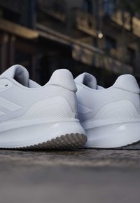 Paar weiße Sportschuhe mit strukturierten Sohlen, die draußen auf rauer Bodenoberfläche liegen, unscharfer urbaner Hintergrund.