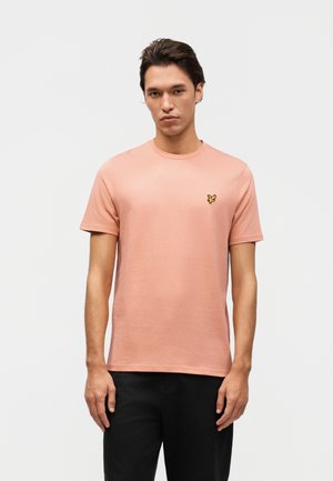 Mladý muž s ružovo lososovým tričkom s krátkym rukávom a malým žltým logom vtáka na ľavej strane hrude, v kombinácii s čiernymi nohavicami.