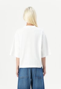 Weißes, übergroßes T-Shirt mit kurzen Ärmeln, Rundhalsausschnitt und lockerer Passform. Kombiniert mit hochgeschnittenen Jeanshosen mit großen Taschen.