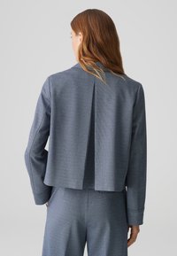Veste structurée bleu avec un pli dans le dos, des manches longues et un tissu texturé. Associée à un pantalon assorti pour un look coordonné.
