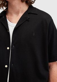 Black short-sleeve shirt with a button-up collar, featuring a subtle logo, light-colored buttons, and a smooth fabric texture.