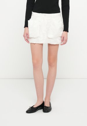 Biała spódnica z kwiatowym wzorem, z dwiema przednimi kieszeniami i prostym krojem. Noszona z czarnymi płaskimi butami, z wstawkami wstążkowymi.