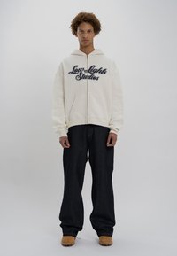 Creme zip-up hoodie med "Low Lights Studios" broderet i marineblå, parret med mørk denim brede benbukser og beige fodtøj.