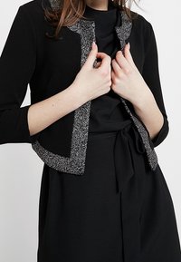 Kvinna som bär en svart klänning med knytliv och en kort svart jacka med silverglittrig trim, som justerar jackkragen med båda händerna.