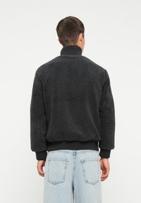 Jachetă neagră din fleece cu un exterior texturat și pufoasă, cu manșete și guler cu nervuri; purtată peste blugi din denim de un albastru deschis.