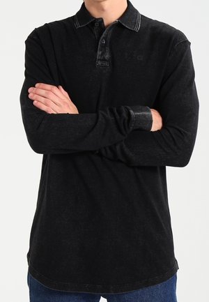 Hombre con camisa polo negra de manga larga con cuello abotonado, brazos cruzados, mostrado desde el cuello hasta la cadera contra un fondo liso.