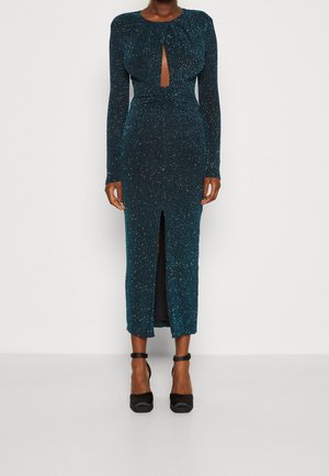 Miętowa, połyskująca sukienka midi z długimi rękawami, wycięciem w kształcie łezki, bocznym rozcięciem i dopasowanym fasonem. Noszona z czarnymi butami na obcasie.