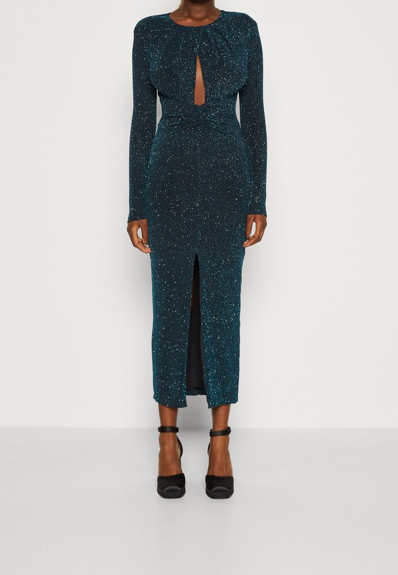 Robe mi-longue pailletée couleur sarcelle, manches longues, découpe en forme de goutte, fente latérale et silhouette ajustée. Portée avec des chaussures à talons noires.
