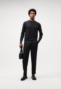 Dark gray sweater, black trousers, and black leather boots. Holds a textured black handbag. Simple, modern design with a relaxed fit.