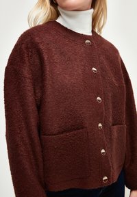 Burgunderfarbener Wollmischungs-Cardigan mit strukturiertem Finish, rundem Ausschnitt, vorderen Taschen und silberfarbigen Knöpfen. Lässige, oversized Passform.