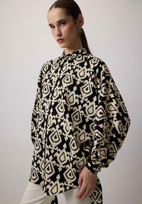 Bluză neagră și ivoire cu un model geometric, cu guler înalt și mâneci voluminoase; confecționată dintr-un material neted cu un finisaj lucios.