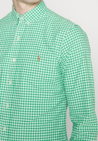 Košile se zeleným a bílým kostkovaným vzorem gingham, s límečkem na knoflíky, malým vyšívaným logem na hrudi a klasickým zapínáním na knoflíky.