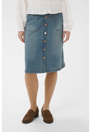 Mujer con falda vaquera azul hasta la rodilla con botones delanteros y bolsillos, combinada con mocasines marrones y una blusa blanca de manga larga.