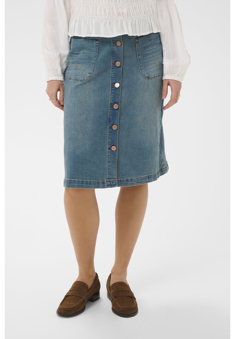 Moteris vilki mėlynos spalvos džinsinę sijoną iki kelių su priekinėmis sagomis ir kišenėmis, derina jį su rudais loaferiais ir balta ilgomis rankovėmis palaidine.