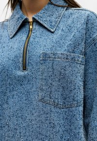 Veste en denim bleu texturé avec une poche poitrine sur le devant et une courte fermeture éclair dorée sous le col.