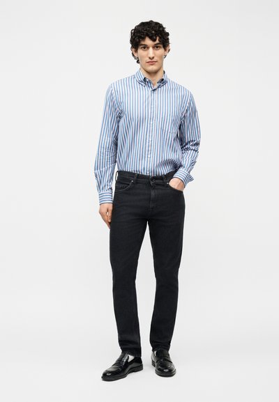 Νεαρός με σγουρά μαλλιά φοράει γαλάζιο και άσπρο ριγέ πουκάμισο με κουμπιά, μαύρο τζιν και μαύρα δερμάτινα παπούτσια, στέκεται με το ένα χέρι στην τσέπη.