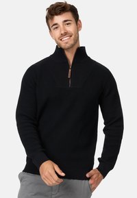 Pull-over en tricot noir avec une fermeture éclair quart, tissu texturé, manches et ourlet côtelés, présentant une petite tirette en cuir marron sur la fermeture éclair.