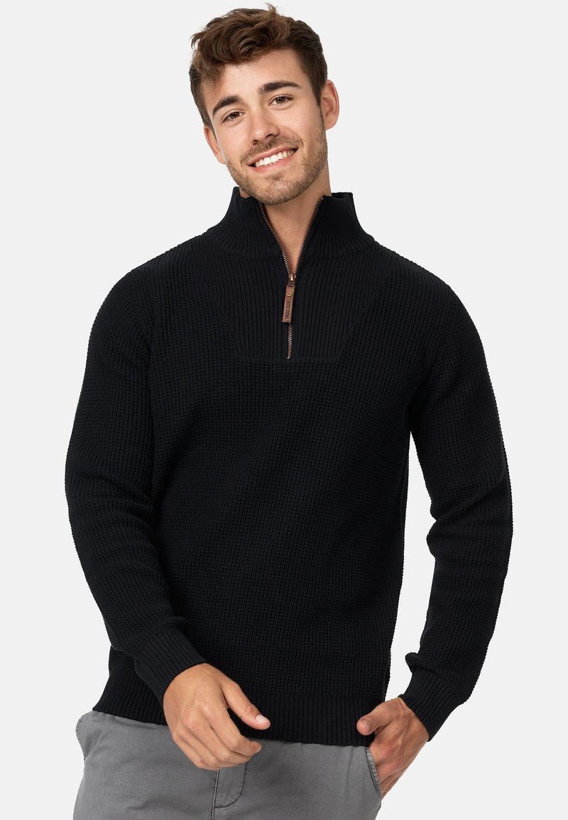 Pull-over en tricot noir avec une fermeture éclair quart, tissu texturé, manches et ourlet côtelés, présentant une petite tirette en cuir marron sur la fermeture éclair.