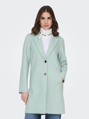 Muntgroene wolmix jas met reverskraag, twee voorzakken en een enkele knoopsluiting. Gecombineerd met een witte, getextureerde sjaal.