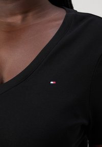 Camiseta negra de cuello en V hecha de una tela suave, con un pequeño logo bordado en colores rojo, blanco y azul en el lado izquierdo del pecho.
