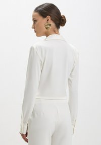 Blouse blanche à manches longues avec col, présentant des poignets boutonnés et un tissu lisse et texturé. Le modèle porte de grandes boucles d'oreilles circulaires en or.