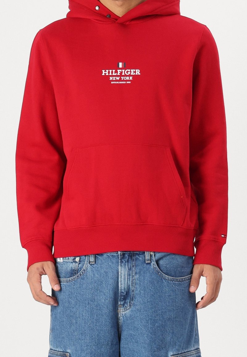 Personne portant un sweat à capuche rouge Hilfiger avec poche avant et un short en jean bleu, debout devant un fond uni.