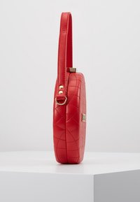 Červená prošívaná kožená kulatá taška s dlouhým popruhem, zdobená zlatými detaily a texturovaným povrchem, vystavená na světlem pozadí.