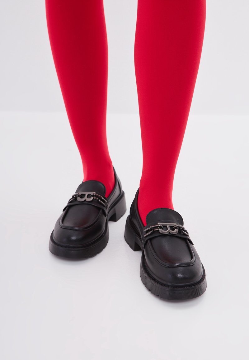 Jambes portant des collants rouge vif et des mocassins en cuir noir avec un détail chaîne argentée, debout sur une surface blanche unie.