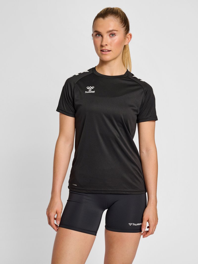 Young woman standing wearing black sportswear: short-sleeve shirt and shorts with white hummel logos on chest and thigh.