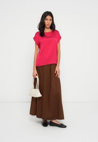 Kobieta stoi przy białej ścianie, ubrana w jaskraworóżowy top, długą brązową spódnicę, czarne baleriny i trzyma małą białą torebkę w lewej ręce.