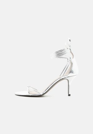Sølvfarget høyhælte sandal med tynne stropper, ankelsnøring og slank stilettohæl på en enkel hvit bakgrunn.