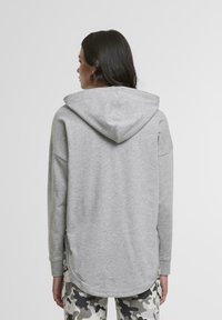 Szary hoodie z zaokrąglonym dołem i dużym kapturem. Miękka tkanina, opuszczone ramiona i długie rękawy. Połączony z spodniami w kamuflaż w wzory.