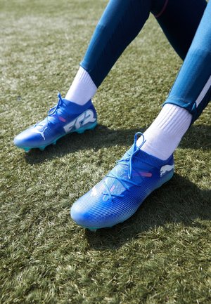 Persoon die blauwe Puma voetbalschoenen, witte sokken en donkerblauwe sportbroek draagt, zit op een grasveld met gestrekte benen.