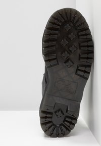 Svart gummisula med djup mönstring, som har en blandning av rundade och kantiga klackar. Sidodetalj visar texturerat material och logotyp.