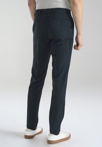 Spodnie slim fit w ciemnym odcieniu teal, wykonane z gładkiego materiału, z kieszeniami z tyłu, noszone z białymi sneakersami, prezentujące smukły design.