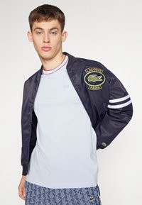 Námořnická bomber bunda s bílými pruhy, vyšívaným logem Lacoste, spárovaná se svetříkem světle modré barvy a vzorovanými šortkami. Hladká textura materiálu.