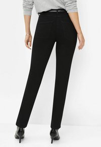 Pantalons noirs à jambe droite avec une texture lisse, deux poches arrière et une taille haute, portés avec des chaussures à talons noires et un haut gris clair.
