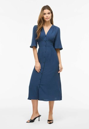 Femme portant une robe midi bleu marine boutonnée avec des manches aux coudes et des sandales noires à talons, debout sur un fond blanc.