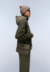 Felpa con cappuccio verde oliva texture, dotata di zip e cappuccio, abbinata a pantaloni verdi oliva. Un berretto marrone chiaro e una borsa a tracolla completano il look.