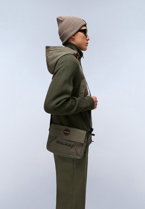 Olivgrüner, strukturierter Kapuzenpullover mit Reißverschluss und Kapuze, kombiniert mit passenden olivfarbenen Hosen. Eine hellbraune Mütze und eine Umhängetasche vervollständigen das Outfit.