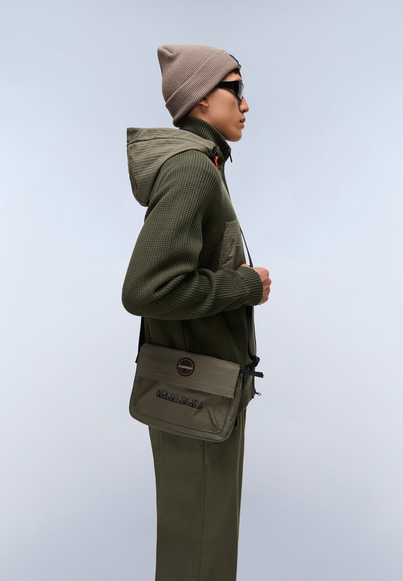 Felpa con cappuccio verde oliva texture, dotata di zip e cappuccio, abbinata a pantaloni verdi oliva. Un berretto marrone chiaro e una borsa a tracolla completano il look.