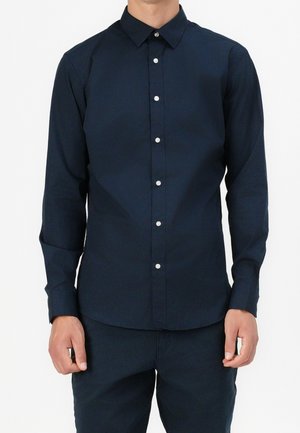 Hombre que lleva una camisa azul marino ajustada de manga larga con botones blancos, de pie frente a un fondo blanco liso.