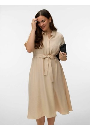 Ženska v bež midi obleki z pasom drži črno torbico, se dotika las in stoji pred enobarvnim belim ozadjem.